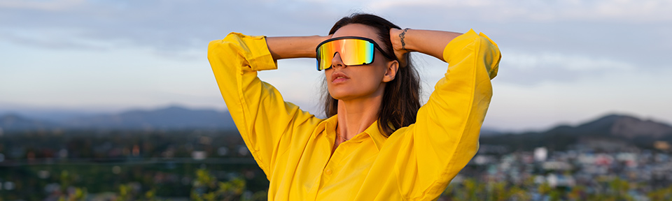 Frau trägt eine reflektierende Sportsonnenbrille und ein gelbes Hemd, während sie vor einer Berglandschaft posiert.