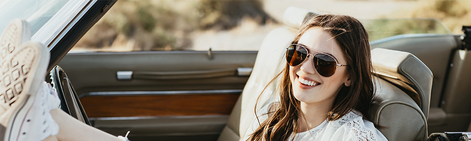 Eine junge Frau sitzts in einem Auto und trägt eine American Optical Sonnenbrille während Sie lächelt.