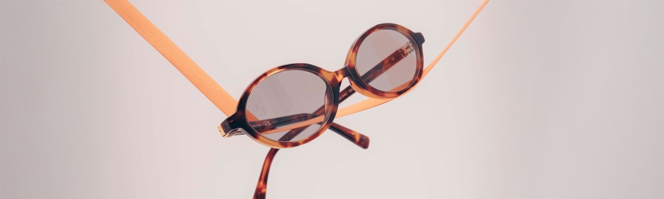 Runde Sonnenbrille mit Schildpattmuster von Miu Miu, die an zwei orangefarbenen Bändern aufgehängt ist, vor einem hellen, neutralen Hintergrund.