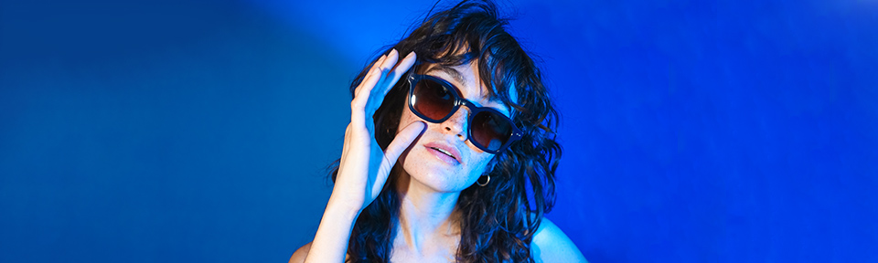 Frau mit lockigem Haar, trägt eine Sonnenbrille und hebt eine Hand vor ihrem Gesicht, vor blauen Hintergrund.