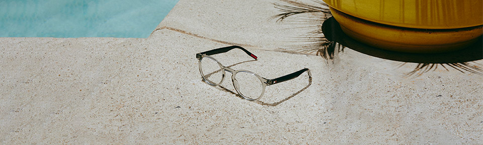 Eine transparente Tommy Hilfiger Brille liegt an einem Pool auf einer Steinfläche.