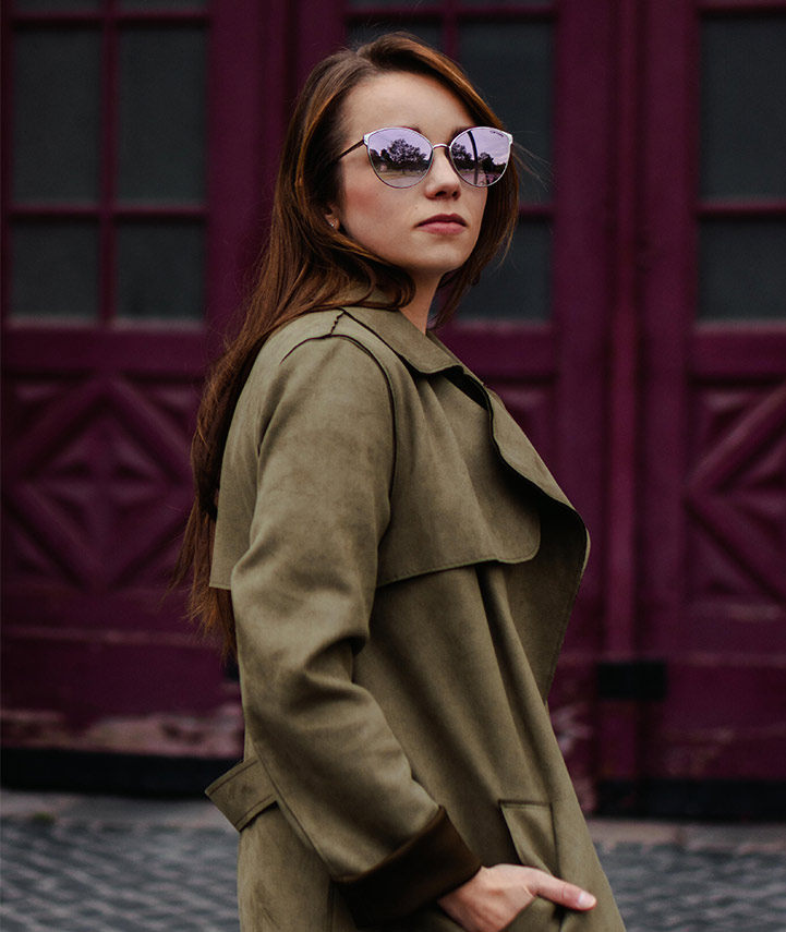 Frau mit rosé-verspiegelter Sonnenbrille und olivgrünem Mantel vor dunkelviolettem Holztor.