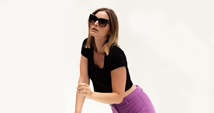 Frau mit schwarzer Sonnenbrille und schwarzem Shirt posiert vor hellem Hintergrund in lila Hose.